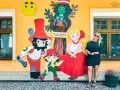 Kobieta pozuje przy kolorowej ścianie Muzeum Zabawek w Kudowie-Zdroju, Dolny Śląsk. Wesołe postacie z bajek towarzyszą jej w radosnej scenerii.