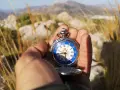 Mano sostiene un reloj de bolsillo antiguo con esfera azul y números romanos, útil para orientarse con reloj de agujas en un paisaje montañoso.