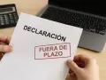 Manos sostienen un documento de "DECLARACIÓN" con un sello rojo que dice "FUERA DE PLAZO". Detrás, una calculadora y un portátil. Pregúntate: ¿qué pasa si no hago la declaración de la renta?