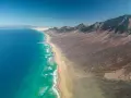 Dzika plaża Cofete z turkusową wodą i surowymi górami w tle. Długi pas piasku oddziela ocean od pustynnego krajobrazu.