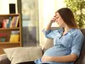 Kobieta w ciąży siedzi na kanapie, trzymając się za czoło. Czuje zawroty głowy w ciąży.