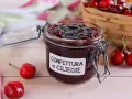 Un vasetto di marmellata di ciliegie fatta in casa, con ciliegie fresche e un cestino sullo sfondo.