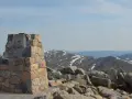 Kamienny monument na szczycie góry Kościuszko, otoczony głazami, z widokiem na ośnieżone pasma górskie w oddali.
