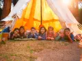 Sept enfants souriants allongés devant une tente de camping éclairée par le soleil.
