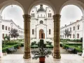 A courtyard with a cross and potted plants, framed by arches. The white church in the background is a place where monks like Father John and Brother Michael find peace.