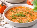 Eine cremige Tomatensuppe mit Reis, garniert mit frischer Petersilie und Basilikum. Daneben liegen frische Tomaten und Gewürzkörner.