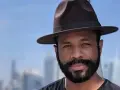 Hombre con sombrero y barba espesa, sonriendo ante un fondo de edificios y cielo azul.