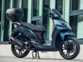 Scooter SYM Symphony SR 125 azul oscuro con baúl trasero, aparcada frente a un edificio moderno.