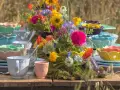 Färgglada uppläggningsfat i plast, staplade med blommor och glas, skapar en festlig dukning utomhus.