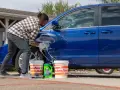 A man washes a blue car in the sun, with buckets and car wash soap nearby. It's a good day to wax a car in the sun.