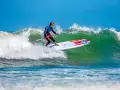 Ein Mann im Neoprenanzug surft auf einem SUP-Board eine grüne Welle. Das SUP-Wellenreiten erfordert Balance und Geschick.