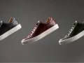 Sneakers marche famose in pelle nera, bordeaux e verde, con suola bianca, fluttuano su sfondo grigio.