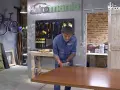 Hombre lijando mesa de madera. El taller tiene herramientas colgadas y una bicicleta morada. La madera hinchada por humedad se repara.