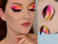 Oczy z wyrazistym makijażem cut crease w odcieniach różu, pomarańczy i żółci. Czarne kreski i długie rzęsy podkreślają spojrzenie.