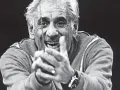 Uśmiechnięty Leonard Bernstein, dyrygent, z dłońmi złożonymi przed sobą, wpatruje się w widza.
