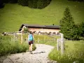 Frau mit Rucksack wandert auf einem Schotterweg zu einer Berghütte. Grüne Wiesen und Bäume im Hintergrund.