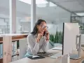 Donna sorridente al telefono in ufficio, mentre lavora al computer. Potrebbe chiedersi quanto guadagna una segretaria al mese.