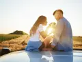 Ein Paar sitzt auf einem Auto im Sonnenuntergang, ein Hund zwischen ihnen. Sie scheinen in einer stillen Partner Meditation zu sein, umgeben von Feldern.