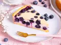 Ein Stück Heidelbeerkuchen Landfrauenart mit saftigen Blaubeeren und cremigem Guss auf einem Teller, daneben eine Gabel und frische Beeren.