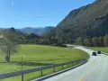 En bil kör på en slingrande väg genom ett grönt landskap med berg och hus. En perfekt start på en semester i Norge.
