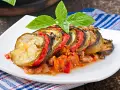 Ratatouille con rodajas de calabacín, berenjena y tomate, decorado con albahaca. Los ingredientes ratatouille se ven tiernos y jugosos.