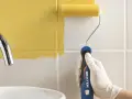 Mano con guante blanco pintando azulejos blancos con rodillo amarillo.