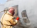Pracownik w kombinezonie i masce wykonuje piaskowanie metalu, przygotowując duży zbiornik do dalszych prac.