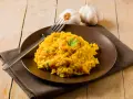 Un invitante risotto zafferano in polvere, guarnito con prezzemolo fresco, su un piatto marrone, con aglio e forchetta sullo sfondo.