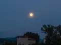 La luna di luglio illumina un paesaggio serale con colline, alberi e un edificio.
