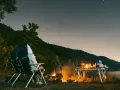 Personne assise près d'un feu de camp sous un ciel étoilé, avec une table de camping et un lac.