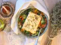 Feta al forno con verdure miste, condita con erbe aromatiche. Accanto, olio, spezie e un mazzetto di origano secco.