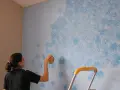 Mujer aplica pintura que disimula imperfecciones en pared con esponja, creando un patrón de nubes azules.