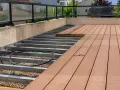 WPC Terrasse Ideen: Unterkonstruktion aus Metall mit Kiesbett und beginnender Verlegung von braunen WPC-Dielen. Moderne Glasbrüstung.