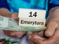 Dłonie trzymają banknoty i drewniane klocki z napisem "14 Emerytura". Tabela emerytura brutto netto jest w tle.