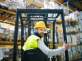 Uśmiechnięty operator wózka widłowego w żółtym kasku i kamizelce odblaskowej, z kciukiem w górze. Zastanawiasz się, ile zarabia operator wózka widłowego?