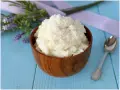 Una ciotola di legno piena di cremosa crema al cocco, guarnita con cocco grattugiato. Accanto, un cucchiaino d'argento e fiori di lavanda.