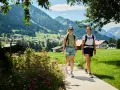 Zwei Frauen wandern durch das Kleinwalsertal mit Blick auf Berge und Häuser. Ein kleiner See ist nicht zu sehen.