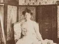 Misia Godebska w wytwornej sukni siedzi na tle bogato zdobionej tapety i parawanu.