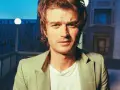 Joe Keery w jasnej marynarce, z naszyjnikiem, na tle nocnego miasta.