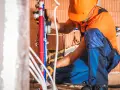 Instalator w pomarańczowym kasku pracuje nad rozprowadzeniem wody w domu, łącząc czerwone i niebieskie rury.