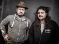 Dwóch mężczyzn, jeden w kapeluszu i z miną figo, drugi z burzą loków, pozuje do zdjęcia.