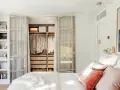 Dormitorio con vestidor integrado. Ropa organizada en armarios abiertos y estanterías. Cama con cojines rosas y manta blanca.
