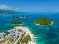 Rajska plaża z zielonymi wysepkami w **jeziorze w Albanii**. Woda jest turkusowa, a na piasku widać leżaki i budynki.