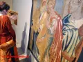 Dwie kobiety oglądają zabytkowy gobelin, przypominający stylem wyczółkowski obrazy. Detale postaci i bogactwo tkaniny przyciągają wzrok.