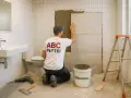 Fachowiec układa płytki na płytki w łazience. Na koszulce widnieje napis "ABC PŁYTKI".