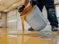 Hombre con guantes vierte pintura para suelo de cemento de color amarillo brillante de un cubo.
