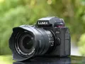 Un appareil photo noir Lumix G9 II avec objectif Leica est posé sur une surface réfléchissante, avec un arrière-plan flou de verdure.