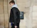 Uomo con un elegante outfit con giubbotto di pelle nero con monogrammi, felpa grigia e pantaloni scuri.