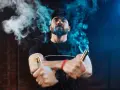 Hombre con barba y gorra, rodeado de vapor azul, sostiene dos vapers, uno dorado y otro cobre. La diferencia entre 0.8 y 1.2 ohm se siente en la calada.