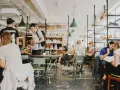 Gente en un café con mesas verdes y estanterías llenas de objetos. El **ruido restaurante** es palpable con las conversaciones y el movimiento.